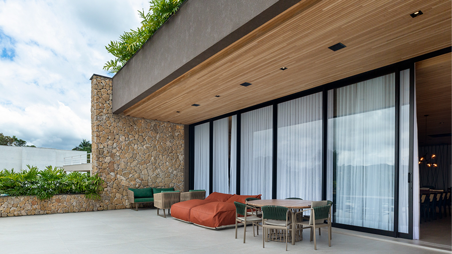 Fachada de casa com lambri de madeira tauari, portas de vidro, parede em pedras e decoração com plantas verdes.