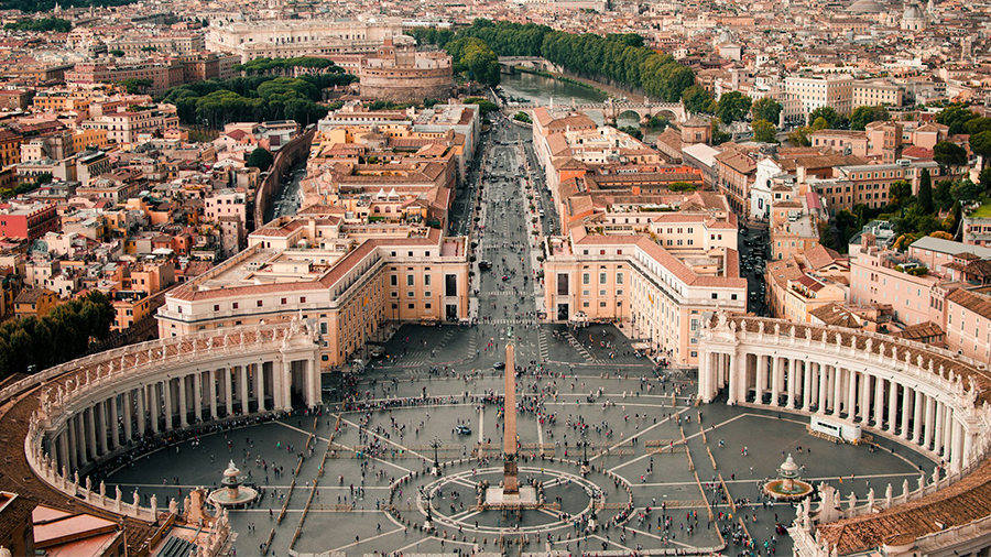  Imagem do Vaticano com o obelisco e construções ao redor formando um semicírculo de cada lado, com a cidade ao redor apresentando diversas casas e prédios.