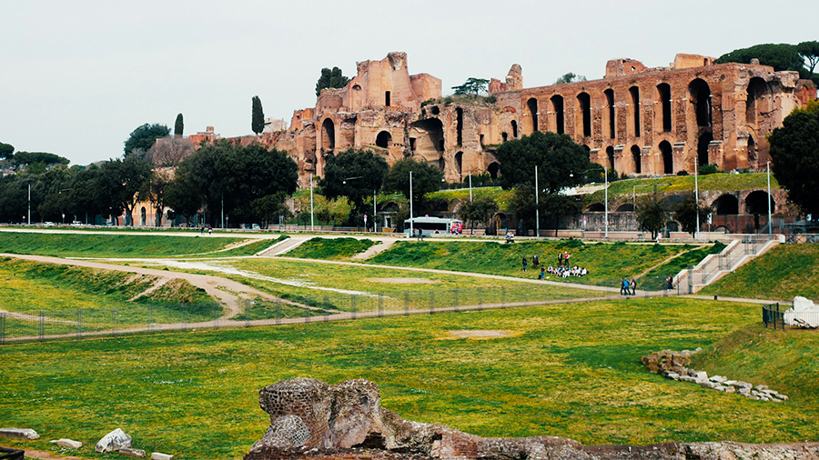 Imagem de uma área com gramados e caminhos, ao fundo uma construção com arquitetura romana e várias árvores ao redor.