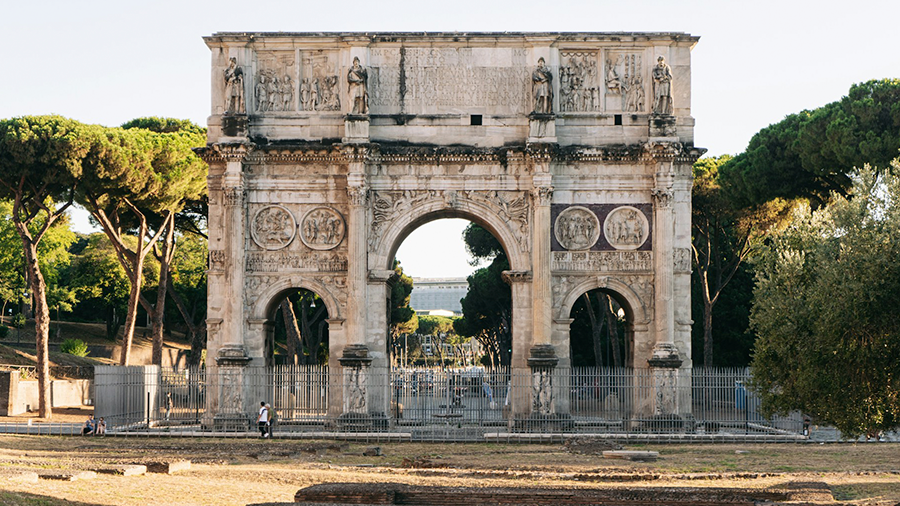 Imagem do Arco de Constantino cercado por grades e árvores ao redor, com pessoas caminhando.