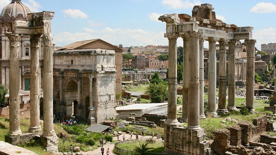 Imagem do Fórum Romano com diversas colunas gregas altas , construções ao fundo, e o ambiente rodeado por grama e caminhos, com pessoas circulando.