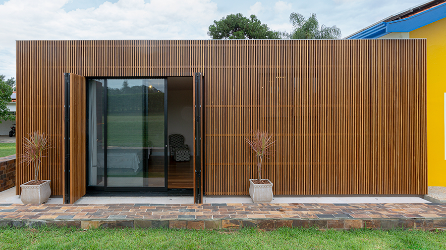 Imagem de uma casa com fachada de vidro e madeira brise vazado ipê, uma porta de vidro e dois vasos de plantas nas laterais. Ao redor, há pisos e grama, e ao lado, uma casa amarela.