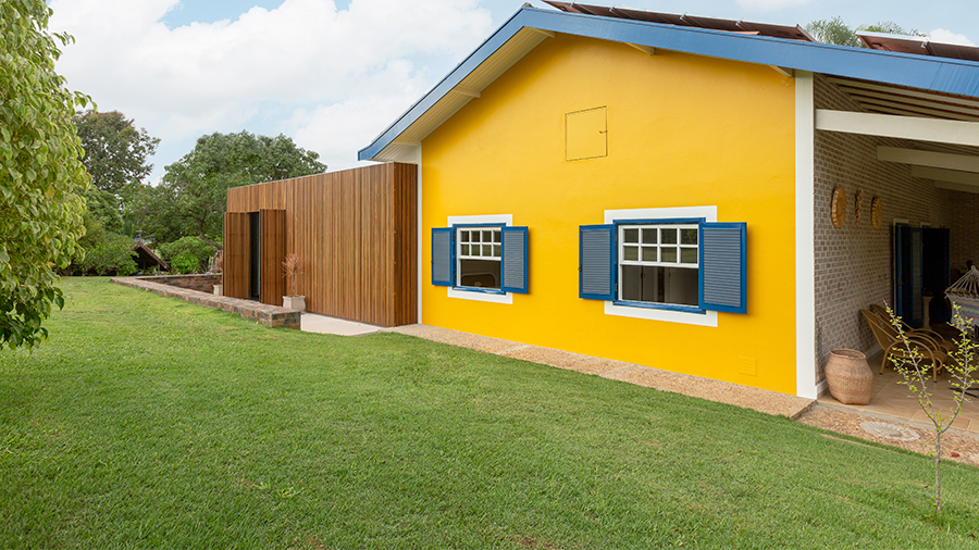  Imagem de um quintal coberto por grama, com uma casa amarela que possui duas janelas e, ao lado, outra casa com fachada de ripas de madeira brise vazado ipê, e árvores ao redor.