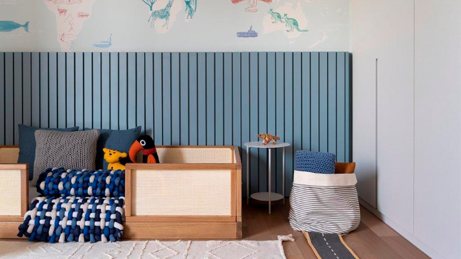 Imagem de um quarto infantil pequeno com uma cama de solteiro com proteção, cobertores e travesseiros. Ao lado, há um saco de brinquedos e uma mesinha. Na parede, há ripas de madeira azuis e um papel de parede com o mapa mundi.