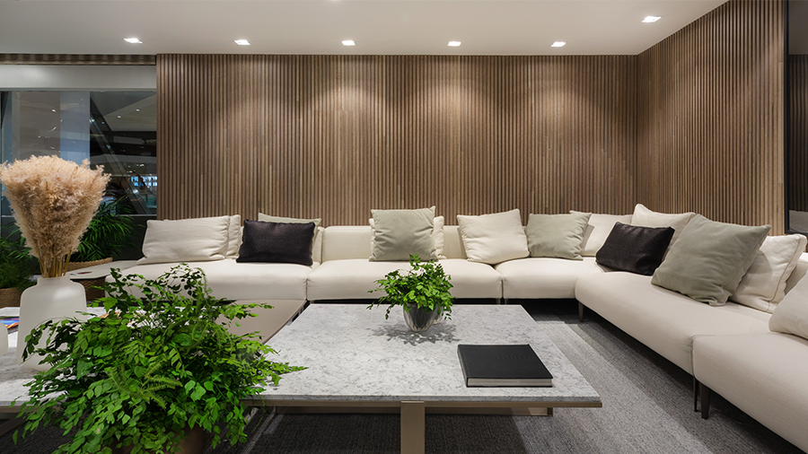  Imagem de uma sala com um sofá grande em L com almofadas, uma mesa de centro com vasos de plantas em cima e na lateral, um tapete, e ao fundo duas paredes com painel ripado de madeira brise tauari com LEDs iluminando do teto.