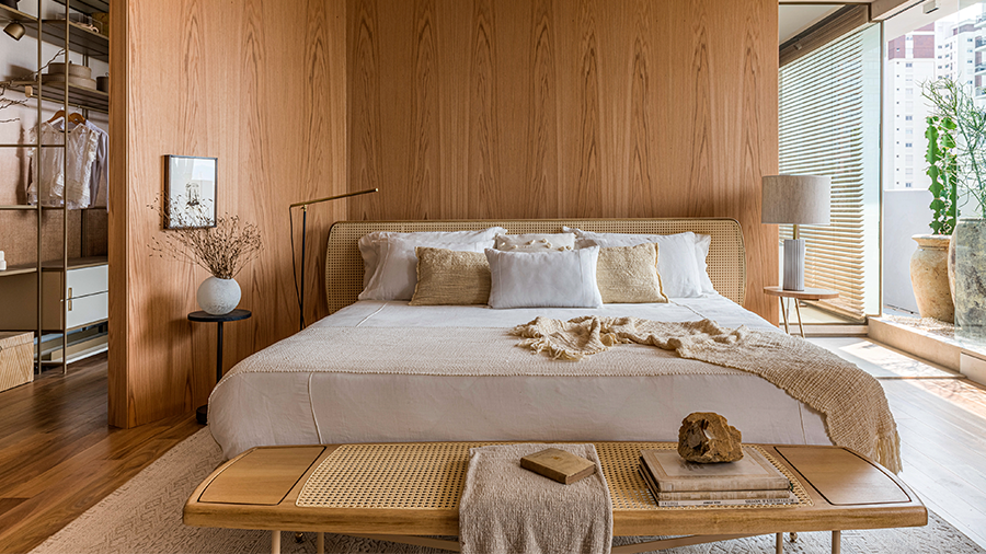 Quarto no estilo japonês, em tons de bege e branco, com uma cama de casal e recamier, parede revestida de madeira e piso de eucalipto citri. Há uma porta de vidro na lateral que dá vista para plantas na área externa.