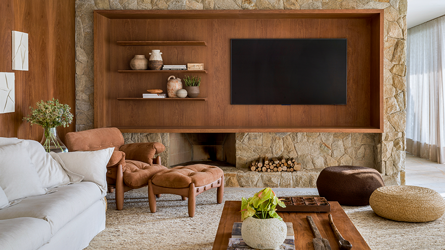 Imagem de uma sala com itens de madeira, um sofá, uma poltrona com um puff na frente, uma mesa no centro e dois puffs ao lado. Ao fundo, uma parede com textura de pedra, um painel de madeira com prateleiras e uma TV ao lado.