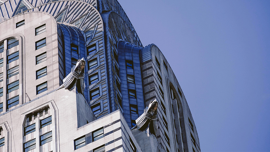 Imagem em close da ponta de um edifício com janelas e formas geométricas, sob um céu azul, destacando a arquitetura art déco.