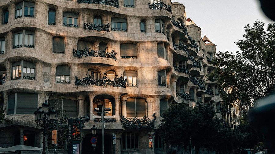 Imagem da Casa Milà que possui arquitetura Art Nouveau vista de frente, com janelas, algumas delas possuem sacadas, postes na frente e árvores ao redor.