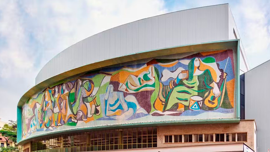 Imagem da fachada curvada do Teatro Cultura Artística, que possui uma faixa com a obra Alegoria das Artes, que possui desenhos coloridos e o céu acima com nuvens.