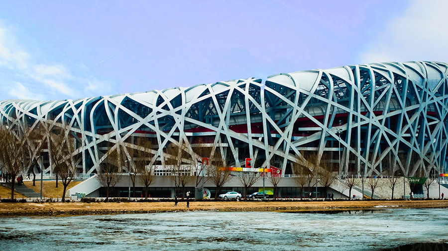 Vista distante do Estádio Nacional de Pequim, com sua arquitetura biomimética e estrutura composta por diversas tiras. Ao redor, há árvores e um lago, sob um céu azul.