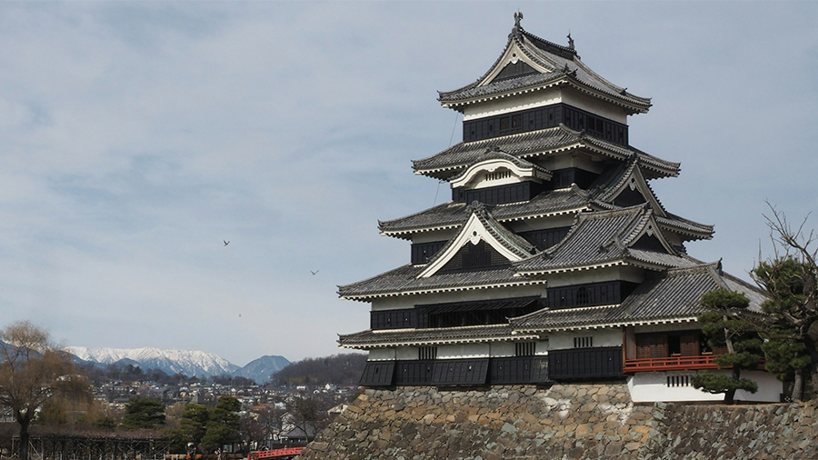 Imagem de uma construção japonesa alta, com arquitetura antiga e cor predominante cinza, cercada por um lago e com um céu repleto de nuvens.