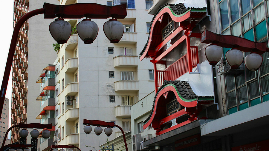 Bairro da Liberdade com prédios de estruturas japonesas em vermelho e preto, cercados por outros edifícios, e lanternas japonesas vermelhas nos postes.