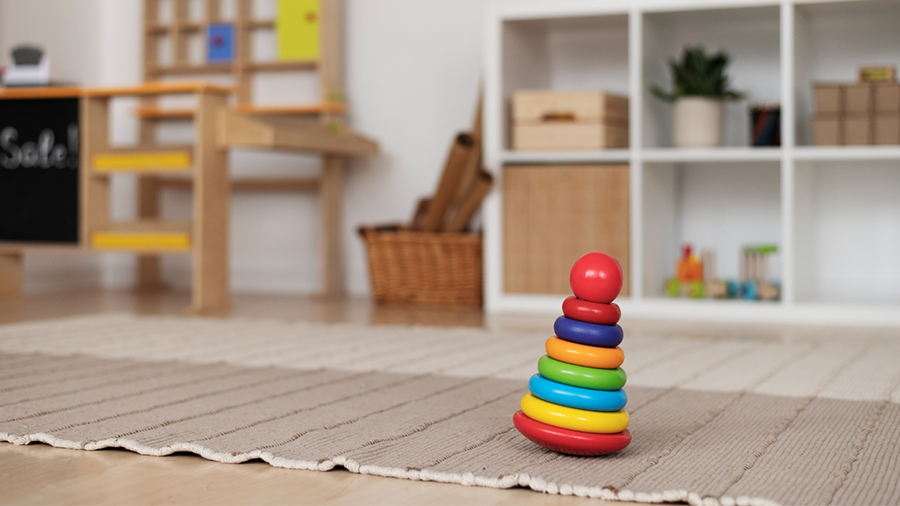 Imagem de uma brinquedoteca no quarto, com um tapete onde há um brinquedo de encaixe colorido. Ao fundo, um armário vazado com objetos e, ao lado, outros brinquedos.