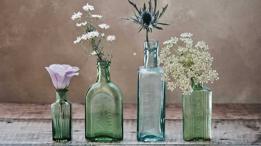 Decoração sustentável com diversas garrafas de formas e tamanhos diferentes, de vidro, com flores de tipos variados em cada uma delas, sobre uma mesa de madeira.