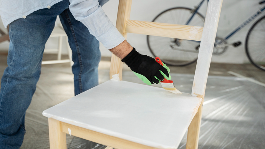 Cadeira usada sendo pintada de branco por uma pessoa de pé ao lado dela, com uma luva e um pincel, e ao fundo, uma bicicleta.